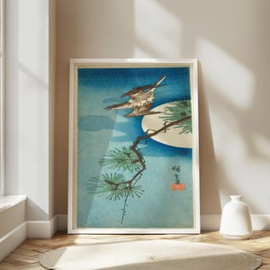 May include: Framed art print featuring a bird in flight above a pine branch, set against a blue background with a full moon. The artwork is in the style of a Japanese woodblock print, with a white frame.