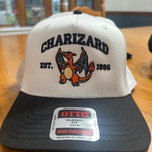 May include: A white baseball cap with a black brim featuring an embroidered graphic of the character Charizard in orange, blue, and yellow. The text "CHARIZARD" is embroidered in black above the character, with "EST. 1996" on the sides. An OTTO brand sticker is on the brim.