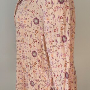 May include: A long-sleeved, pink blouse with a floral print. The print features a variety of flowers and leaves in shades of purple, orange, and brown.