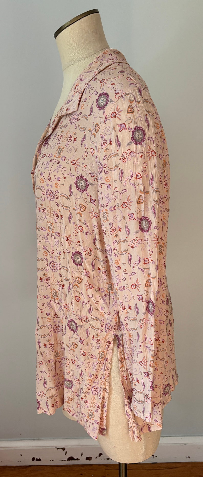 May include: A long-sleeved, pink blouse with a floral print. The print features a variety of flowers and leaves in shades of purple, orange, and brown.
