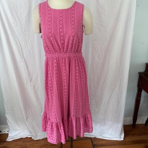 May include: A sleeveless, knee-length pink dress featuring an all-over eyelet lace pattern with a round neckline, a defined waistline, and a ruffled hem. The garment is constructed from a perforated fabric that creates a textured, repeating design.