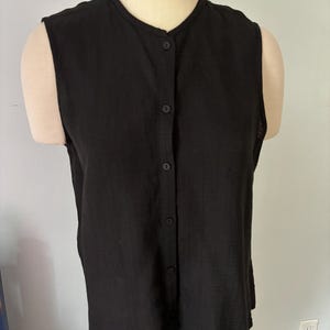 May include: A sleeveless black top with a button-down front. The top has a round neckline and is made of a lightweight fabric. The garment is displayed on a mannequin.