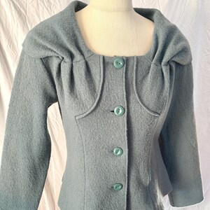 May include: A light blue jacket with a unique square neckline and matching buttons. The jacket has long sleeves and a tailored fit, showcasing a vintage style. The fabric appears to be a textured wool or wool blend.