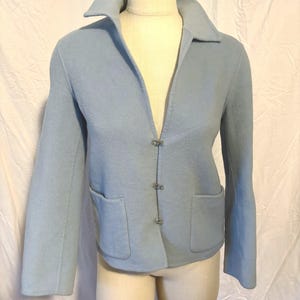 May include: A light blue jacket with a wide collar, long sleeves, and two front pockets. The jacket has three decorative clasps down the front. The jacket is displayed on a mannequin.
