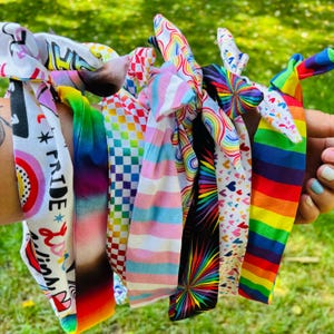 May include: A collection of colorful fabric headbands with various patterns, including rainbow stripes, hearts, and checkered designs. One headband features the word "PRIDE" with a rainbow and other text. The headbands are held up against a blurred green background.
