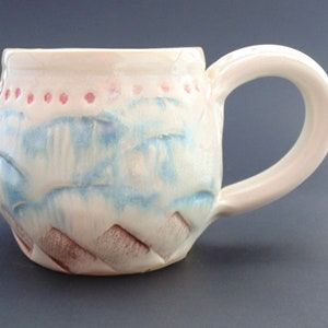 Puede incluir: Una taza de cerámica blanca con un esmalte azul y rosa. La taza tiene un diseño texturizado y una forma redondeada.