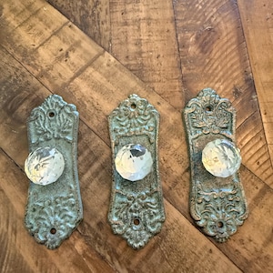 May include: Three antique-style door knobs with clear crystal-like spheres and ornate, weathered, teal-colored backplates. The knobs are arranged on a rustic, wooden surface, showcasing their decorative design.