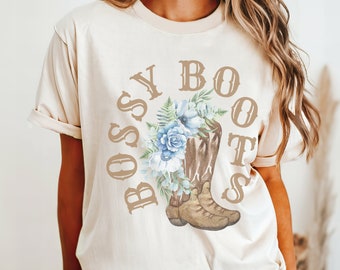 May include: A beige t-shirt with a brown graphic of cowboy boots with blue flowers and the text "Bossy Boots".