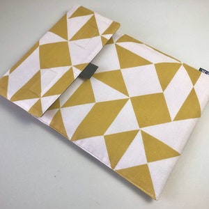 Puede incluir: Una funda rectangular para portátil con un patrón geométrico blanco y dorado. La funda tiene un cierre de solapa y una pequeña etiqueta gris oscuro. El patrón consiste en triángulos y cuadrados. El nombre de la marca está impreso.