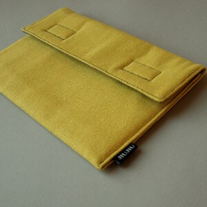 May include: A mustard yellow fabric sleeve with a fold-over flap secured by two rectangular stitched accents. The sleeve features a small black fabric tag on the side with the white text "airate" printed in a lowercase sans-serif font.