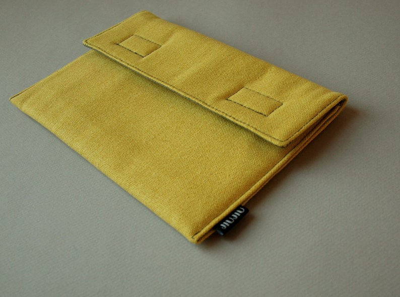 May include: A mustard yellow fabric sleeve with a fold-over flap secured by two rectangular stitched accents. The sleeve features a small black fabric tag on the side with the white text "airate" printed in a lowercase sans-serif font.