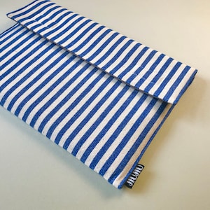 Puede incluir: Una funda o estuche de tela a rayas azules y blancas, posiblemente para un portátil o una tableta. La funda tiene un cierre de solapa y una pequeña etiqueta negra con texto blanco. Las rayas están espaciadas uniformemente y corren horizontalmente por la tela.