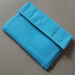 May include: A turquoise fabric case with a flap closure. The case is rectangular with a textured surface and a folded flap secured by three stitched rectangles. The case is on a neutral background.
