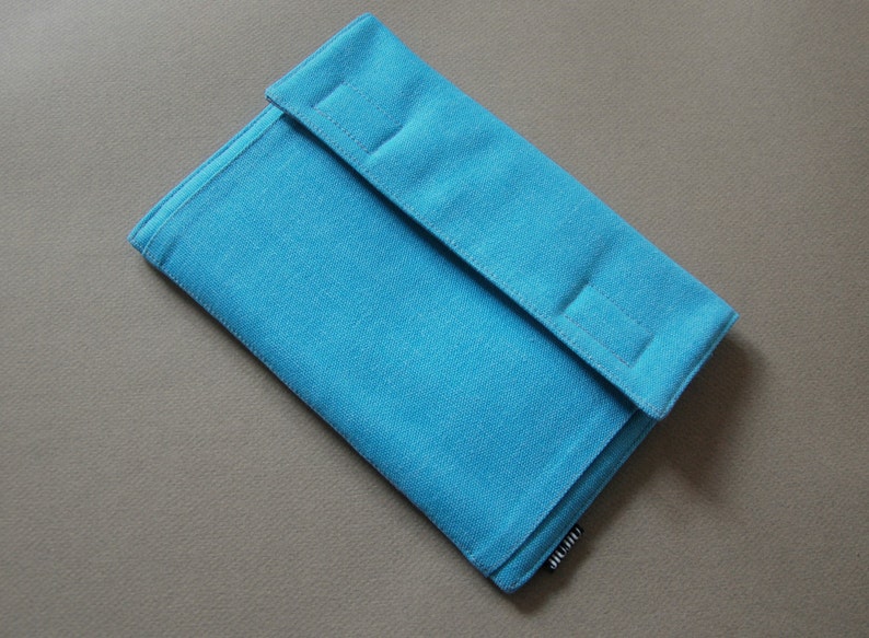 May include: A turquoise fabric case with a flap closure. The case is rectangular with a textured surface and a folded flap secured by three stitched rectangles. The case is on a neutral background.