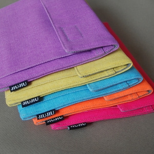 May include: A stack of colorful fabric wallets in shades of purple, yellow, turquoise, orange, and pink. Each wallet has a rectangular shape with a flap and a small black tag that reads "JIJIU". The wallets are made of a textured fabric.