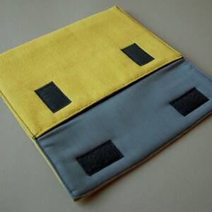 May include: A rectangular fabric pouch featuring a mustard yellow top flap and a slate gray lower section. The design includes four black hook-and-loop fastener strips for closure and a small black fabric tag with white text on the left side edge.