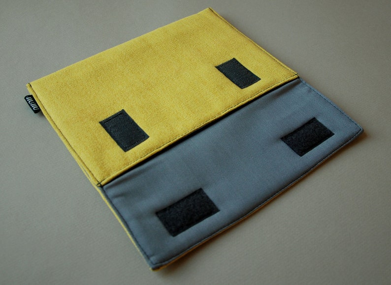 May include: A rectangular fabric pouch featuring a mustard yellow top flap and a slate gray lower section. The design includes four black hook-and-loop fastener strips for closure and a small black fabric tag with white text on the left side edge.
