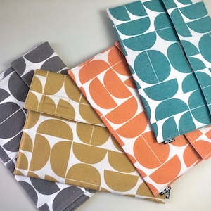 Pode incluir: Uma coleção de capas de tecido em várias cores, incluindo cinza, amarelo mostarda, coral e verde-azulado. Cada capa apresenta um padrão geométrico semicircular branco. As capas são fechadas com uma aba e provavelmente são projetadas para transportar itens.