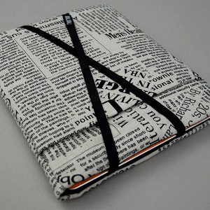 Pode incluir: Estojo coberto de tecido com um design de jornal, preso por uma faixa elástica preta. O estojo, com texto visível, é provavelmente para um tablet ou item semelhante.