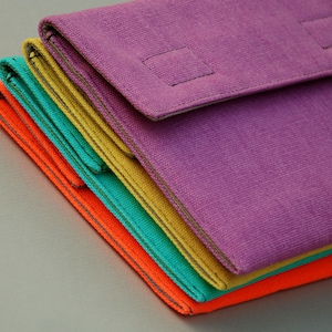 May include: A stack of four rectangular fabric pouches in vibrant colors including purple, yellow, teal, and orange. Each pouch features a fold-over flap with a square stitched detail and visible seams along the edges, creating a layered appearance.