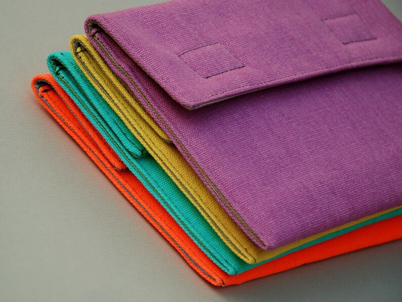 May include: A stack of four rectangular fabric pouches in vibrant colors including purple, yellow, teal, and orange. Each pouch features a fold-over flap with a square stitched detail and visible seams along the edges, creating a layered appearance.