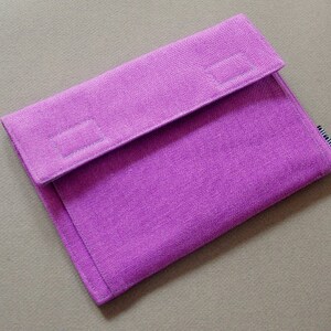 May include: A rectangular purple fabric sleeve with a textured weave and a fold-over flap closure. The flap features two rectangular stitched sections, and a small black and white tag is visible on the right side edge of the sleeve.