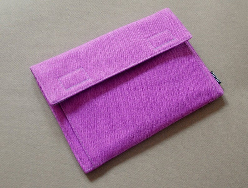 May include: A rectangular purple fabric sleeve with a textured weave and a fold-over flap closure. The flap features two rectangular stitched sections, and a small black and white tag is visible on the right side edge of the sleeve.