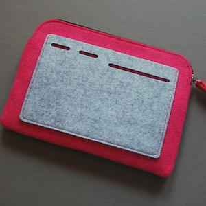 Puede incluir: Una funda con cremallera de fieltro rosa brillante con un bolsillo de fieltro gris cosido en la parte delantera. El bolsillo gris tiene tres pequeños cortes horizontales y un corte largo. El tirador de la cremallera es rosa.