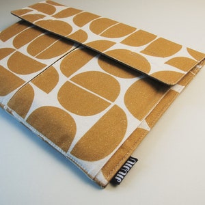 May include: A rectangular, fabric case with a flap closure. The case features a repeating pattern of gold-colored, semi-circular shapes on a white background. A small black tag with white text is visible on the lower right corner.