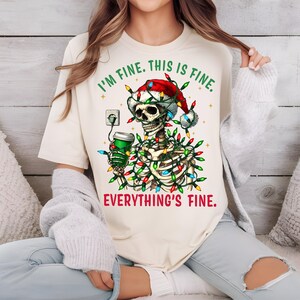 Puede incluir: Camiseta color crema con un esqueleto que lleva un gorro de Papá Noel y luces navideñas. El esqueleto sostiene una taza de café verde y está conectado a una toma de corriente. El texto en la camiseta dice "I'M FINE. THIS IS FINE. EVERYTHING'S FINE."