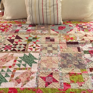 May include: A patchwork quilt with a variety of floral and geometric patterns in shades of pink, green, and brown. The quilt is displayed on a bed with white pillows and a striped pillow.