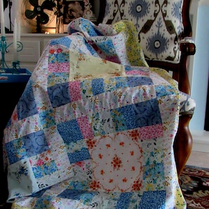 May include: A patchwork quilt featuring a mix of floral and geometric designs in shades of blue, pink, yellow, and white. The quilt is draped over a chair with patterned fabric upholstery.