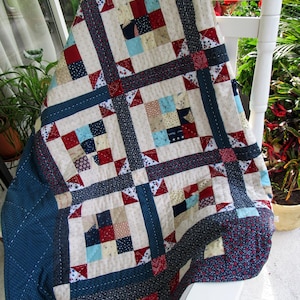 May include: A handmade quilt with a patchwork design in shades of cream, navy blue, red, and light blue. The quilt is draped over a white rocking chair, showcasing its intricate patterns and textures. The quilt features squares and triangles.