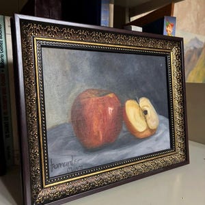Puede incluir: Una pintura al óleo sobre lienzo que muestra una manzana roja entera y una mitad de manzana cortada sobre una superficie gris. La obra está firmada por el artista en la esquina inferior izquierda y se presenta en un marco rectangular ornamentado.