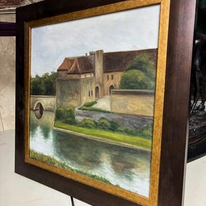 Puede incluir: Pintura al óleo de un castillo con foso, enmarcada en un marco de madera marrón oscuro con un borde interior dorado. La pintura representa un castillo de piedra con un tejado de tejas rojas, un puente y exuberante vegetación. El agua refleja el castillo.