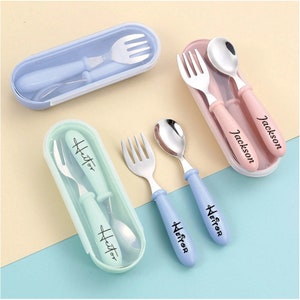 May include: A set of children's stainless steel forks and spoons with colored handles and carrying cases. The handles are blue, pink, and mint green, with names printed on them. The cases are also colored to match the handles.