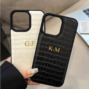 Puede incluir: Dos fundas de teléfono personalizadas. Una es blanca con ribete negro e iniciales doradas "G.F.", la otra es negra con iniciales doradas "K.M.". Ambas fundas tienen un patrón texturizado similar al de la piel de cocodrilo.