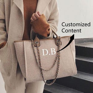 Puede incluir: Un bolso tote beige con asas y ribetes de cuero marrón, con una correa de cadena. El bolso tiene las iniciales "D.B" en blanco. La imagen también incluye el texto "Customized Content".