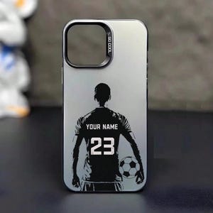 Op de afbeelding: Een zilveren en zwarte telefoonhoes met een voetballerontwerp. Het shirt van de speler toont "YOUR NAME" en nummer 23. De hoes heeft een zilveren frame en de woorden "SO COOL" staan op de zijkant.