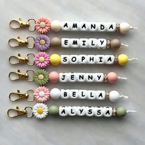Può includere: Sei portachiavi personalizzati con nomi scritti con perline di lettere bianche. Ogni portachiavi presenta un fiore di colore diverso, una chiusura color oro e perline decorative. I nomi includono Amanda, Emily, Sophia, Jenny, Bella e Alyssa.