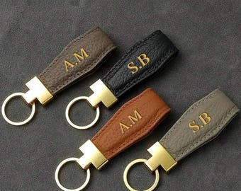 May include: Four leather keyrings in brown, black, tan, and grey, each with gold-coloured hardware and gold-embossed initials. The keyrings are arranged on a dark grey surface.
