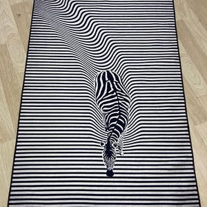 May include: Black and white striped rug with a zebra design. The zebra is facing down and appears to be walking into the stripes.