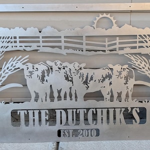 May include: A metal sign featuring a farm scene with cows, a fence, and wheat. The sign includes the text "THE DUTCHIK'S EST. 2010". The design is cut out of the metal, creating a silhouette effect. The sun is visible in the background.