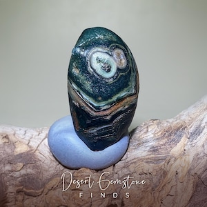 May include: A polished, oval-shaped gemstone with a dark green base, featuring concentric patterns of white, brown, and black. The stone rests on a light blue, rounded base, with the text "Desert Gemstone FINDS" at the bottom.