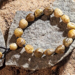 May include: A beaded bracelet made of smooth, rounded, light brown and tan stones. The bracelet is strung on a black cord and rests on a gray rock, with a sandy background. The stones vary in size and have a natural, organic appearance.