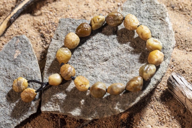 May include: A beaded bracelet made of smooth, rounded, light brown and tan stones. The bracelet is strung on a black cord and rests on a gray rock, with a sandy background. The stones vary in size and have a natural, organic appearance.