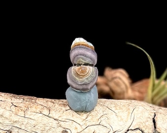 May include: A small, stacked stone sculpture. The sculpture is composed of four stones in shades of blue, purple, tan, and white. The stones are stacked on top of each other, creating a balanced and artistic arrangement. The sculpture is set against a dark background.