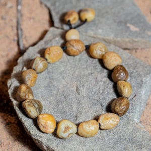 May include: A handmade bracelet featuring smooth, rounded, beige and brown stones strung on a black cord. The bracelet is displayed on a gray stone, with a sandy background and a twig.