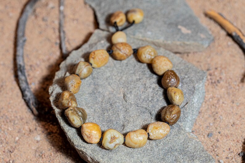 May include: A handmade bracelet featuring smooth, rounded, beige and brown stones strung on a black cord. The bracelet is displayed on a gray stone, with a sandy background and a twig.