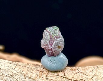 May include: A small, polished, multi-layered stone featuring swirling patterns of pink, white, and pale green. The stone is mounted on a smooth, rounded, light blue base, creating a distinct contrast between the intricate textures and the solid color.